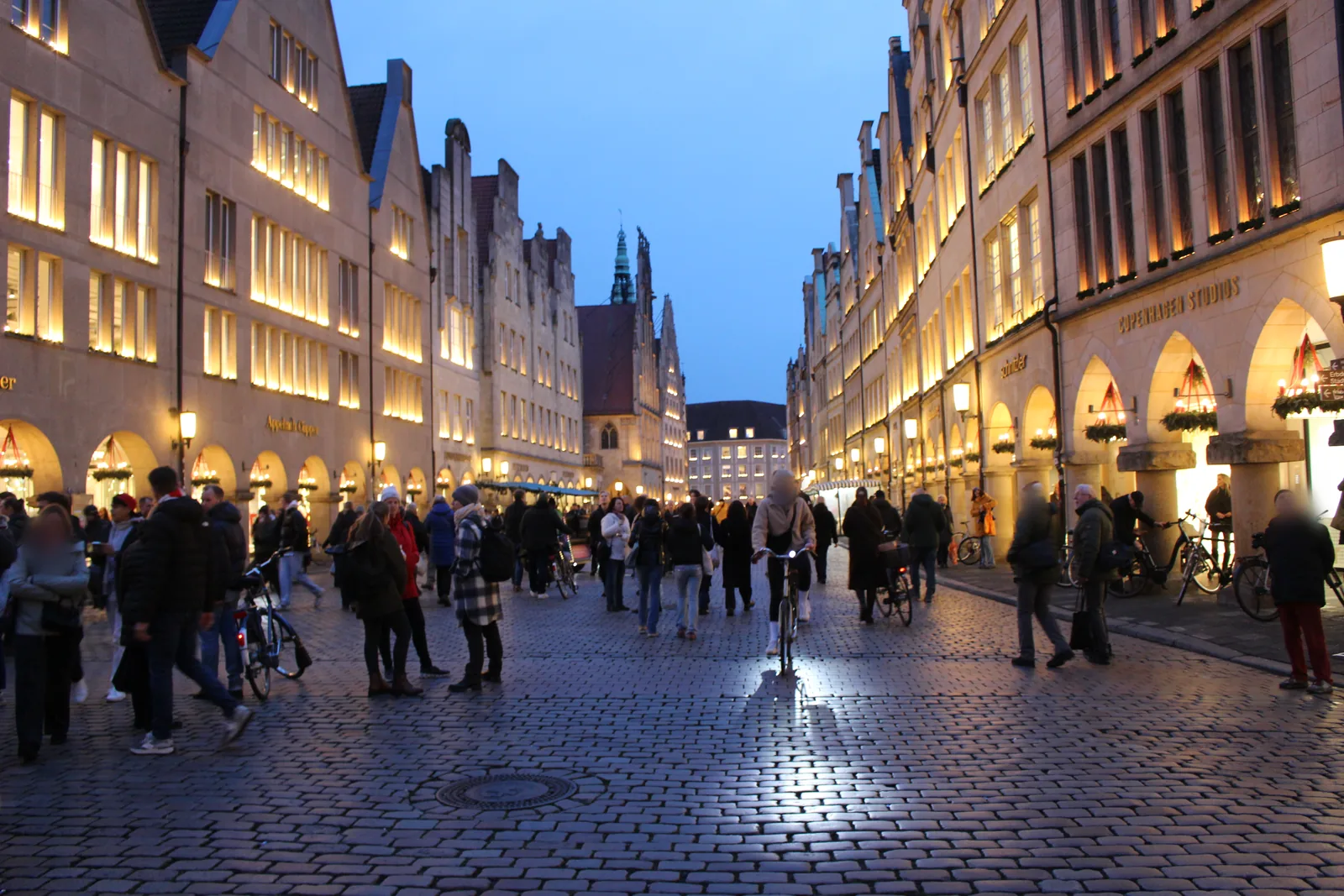 Prinzipalmarkt in Münster at blue hour: warmly lit gabled houses and arcades with advent wreaths, people walking and cycling on the rain-wet cobblestones.