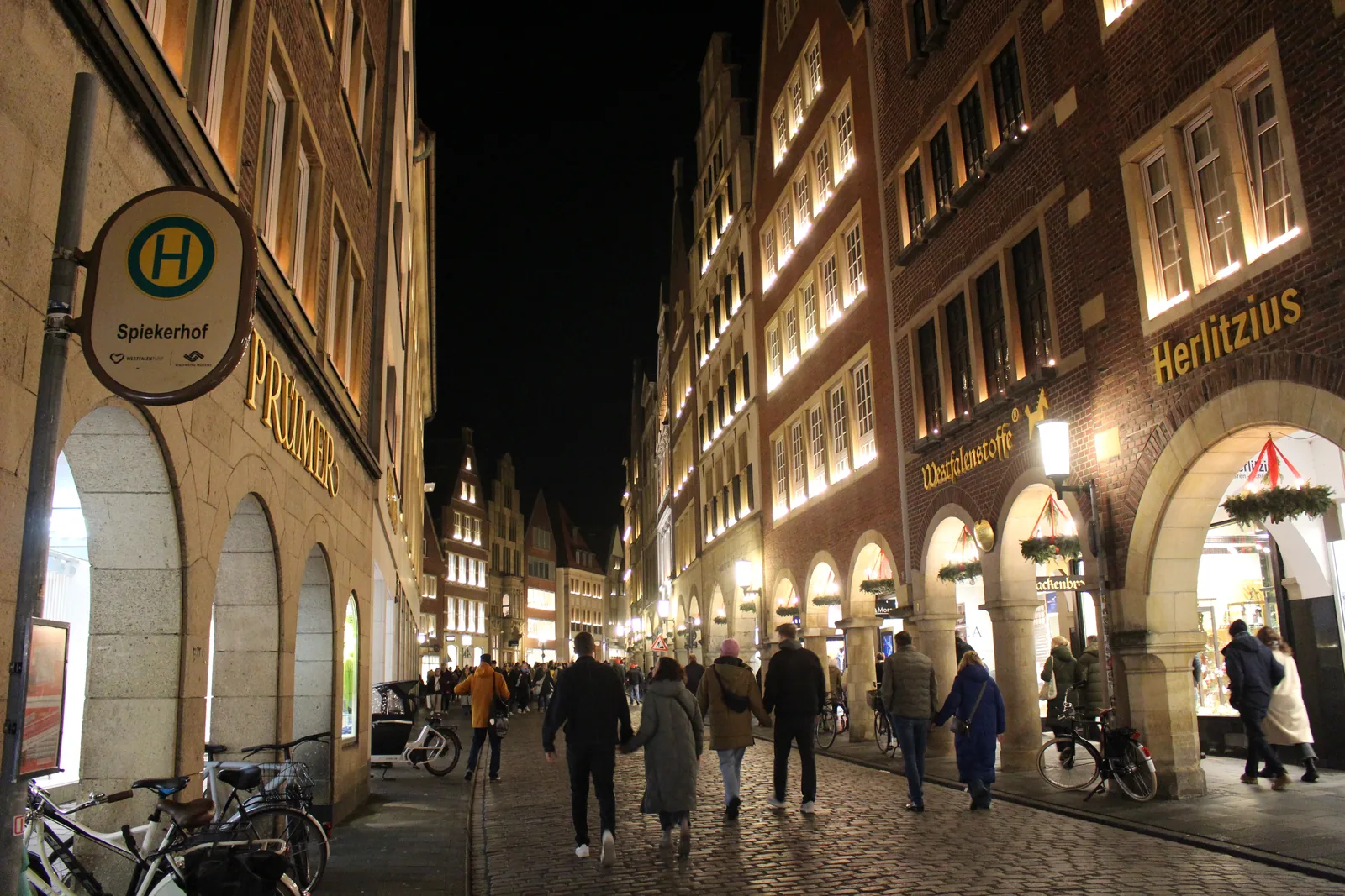 Gabled houses and arcades with advent wreaths lit up in the evening on the Roggenmarkt in Münster, people strolling on the cobblestones, the Spiekerhof bus stop sign in front.