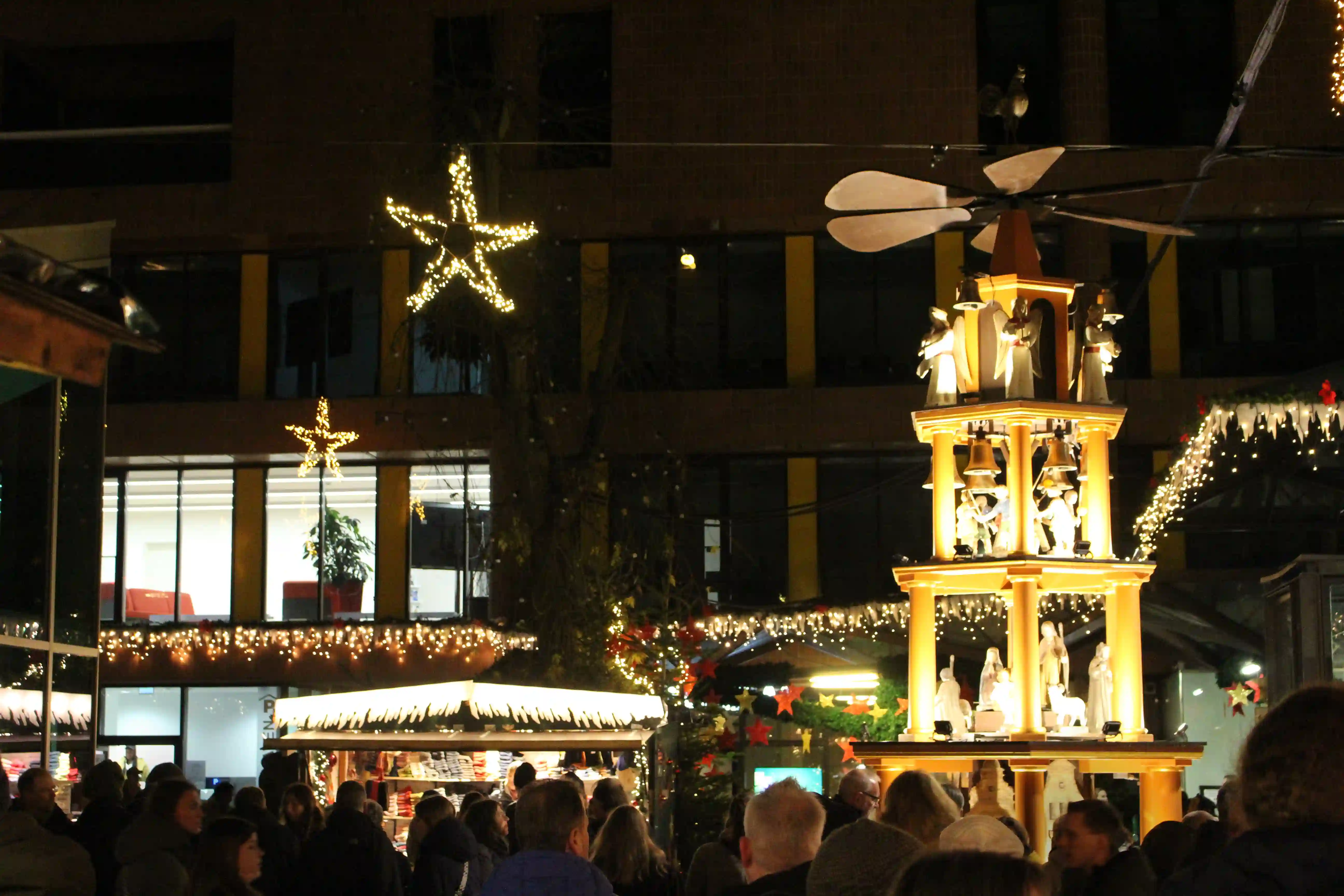 Large lit wooden pyramid with vane wheel, angels and bells at the Aegidii Christmas market in Münster, above the visitors in the courtyard.
