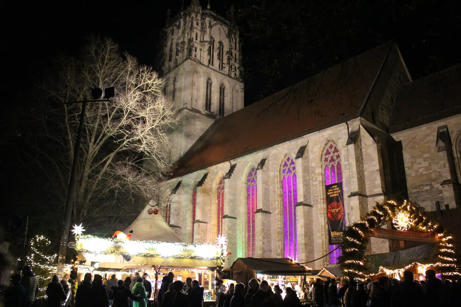 Giebelhüüskesmarkt in front of the colourfully lit Liebfrauen-Überwasser church in Münster in the evening, with stalls and people.
