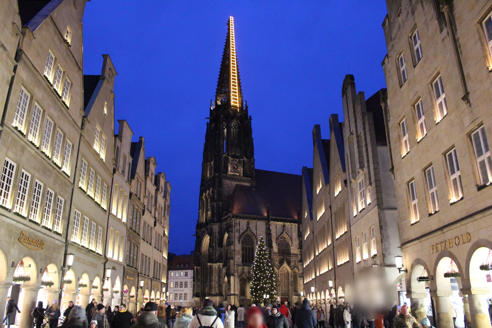 Prinzipalmarkt in Münster at blue hour: the gold-lit Himmelsleiter installation on the tower of St. Lamberti's above the Christmas market and the large Christmas tree.